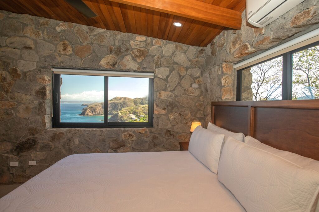 Guest bedroom with a white bed, stone walls, wood ceiling and a window overlooking Ocotal Bay