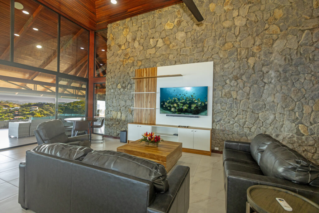 Black leather sofas and wall-mounted TV in the stone-walled living room, with the veranda beyond